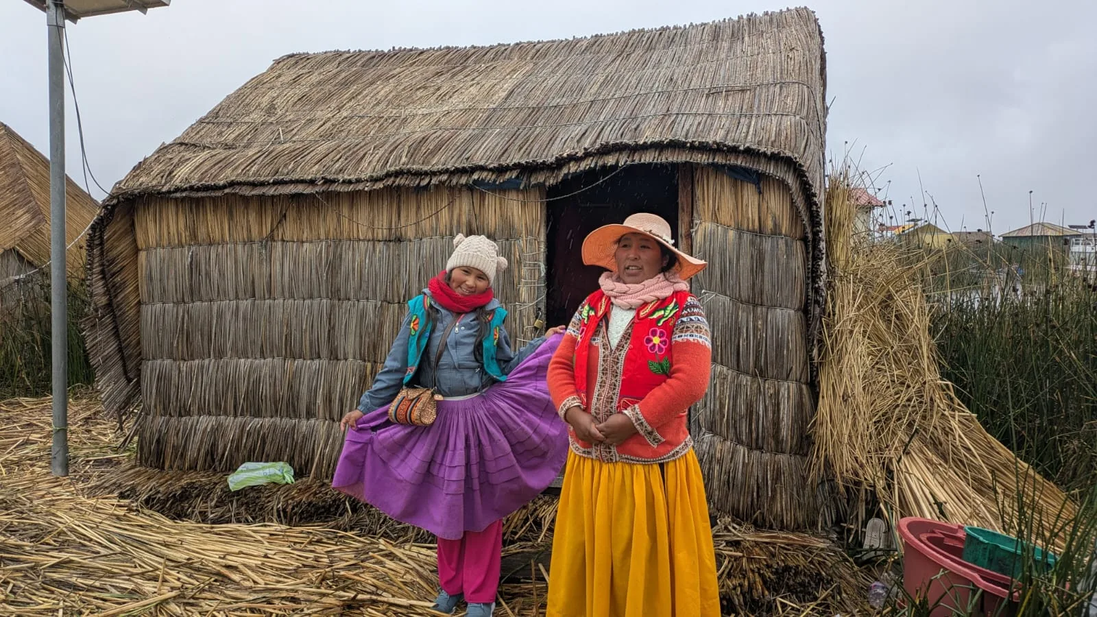 Dos mujeres con ropa colorida conversan junto a una vivienda tradicional de totora